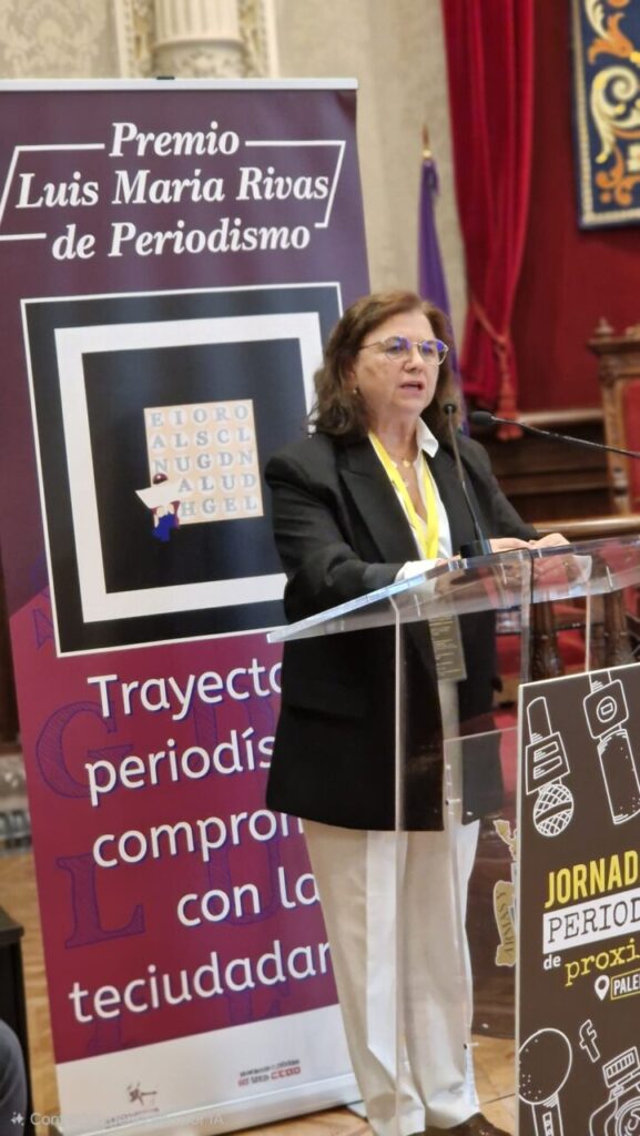 Mujer hablando en un evento sobre periodismo con un cartel detrás.