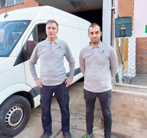 Dos hombres de pie frente a una furgoneta blanca en un taller de reciclaje