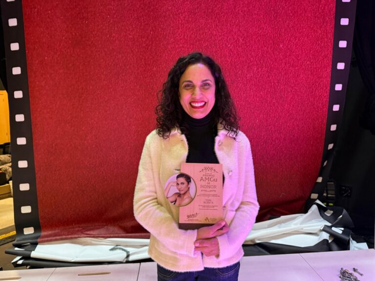 Toni Acosta sonriente sosteniendo el Premio AMGu de Honor en un escenario.