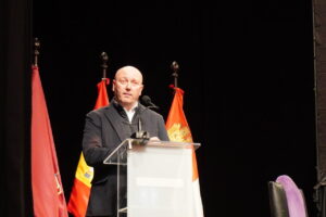 Hombre hablando en un evento con banderas de fondo