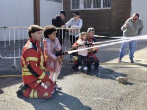 Niños usando mangueras con bomberos en jornada de puertas abiertas