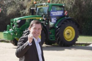 Candidato del PP sonriendo frente a un tractor John Deere en campaña electoral.