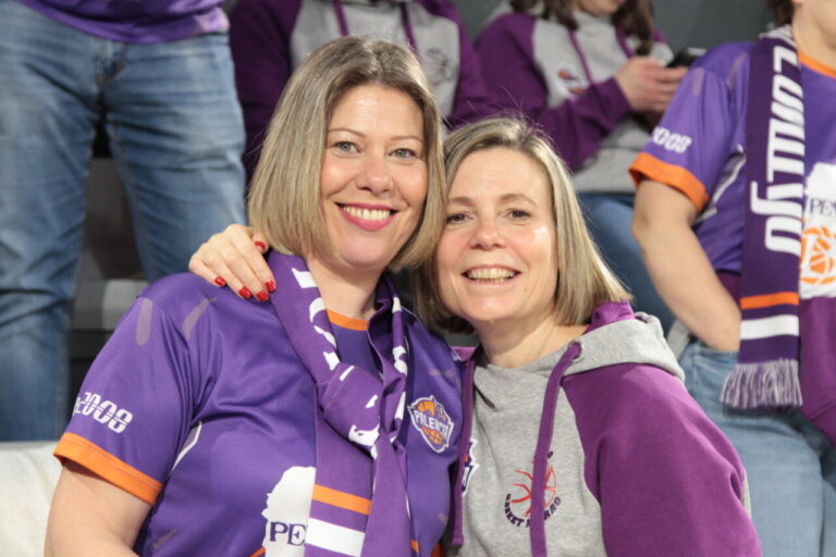 Dos aficionadas sonrientes con camisetas moradas en un partido de baloncesto.