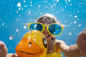 Niña sonriendo con gafas grandes y un inflable de pato en el agua