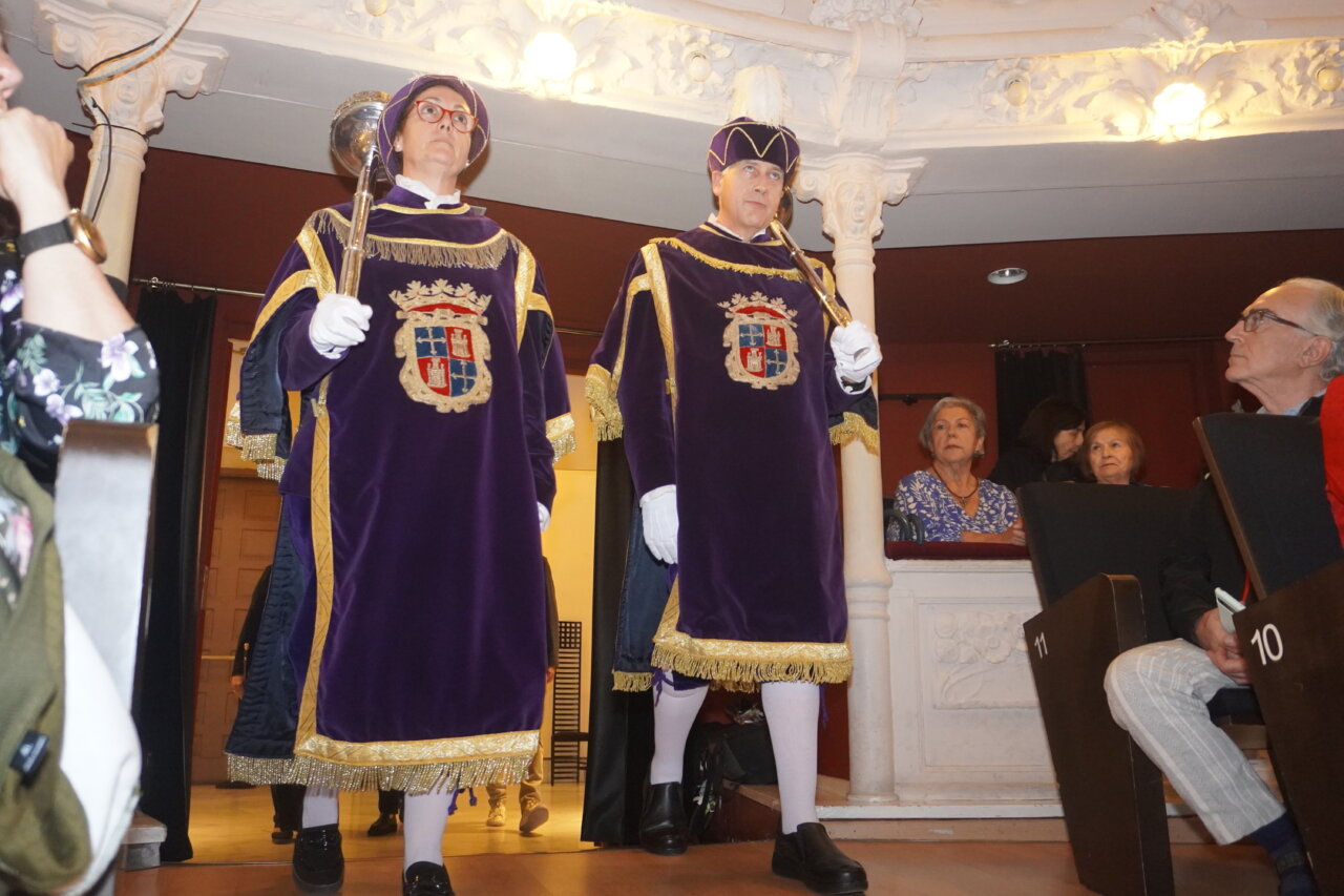 Dos personas vestidas con trajes ceremoniales en un teatro durante el pregón literario.