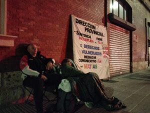Dos personas sentadas en sillas frente a un cartel de protesta