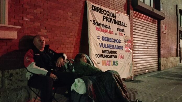 Dos personas sentadas en sillas frente a un cartel de protesta
