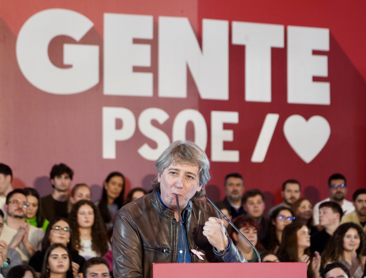 Un hombre habla ante un micrófono frente a asistentes y un fondo con el lema PSOE.