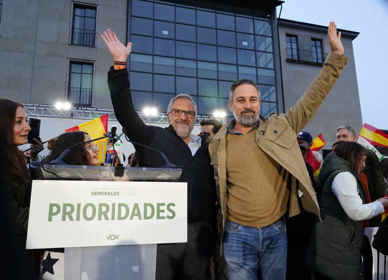 Dos hombres saludan con la mano en alto junto a un atril de Vox.