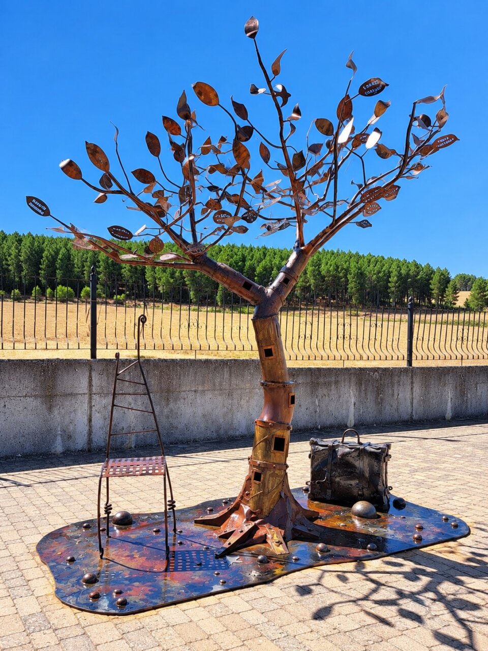Escultura de hierro con forma de árbol y silla metálica en un patio soleado.