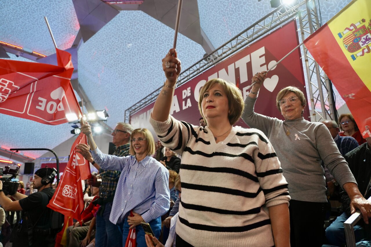 Asistentes a un mitin levantan banderas rojas bajo una pancarta política.
