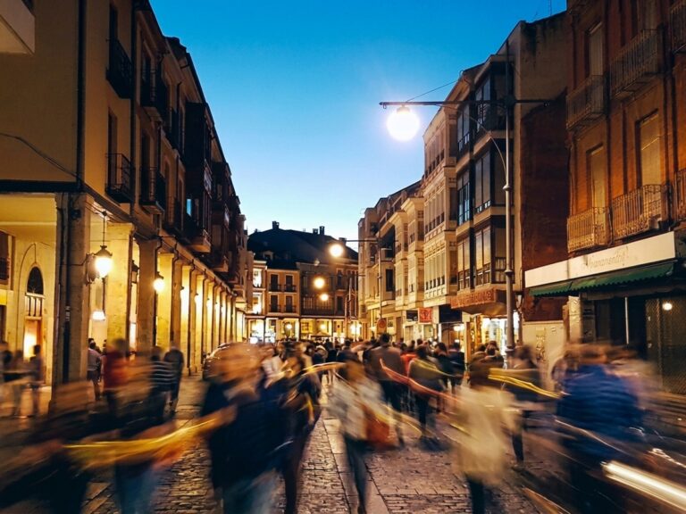 Calle peatonal iluminada al anochecer, con edificios comerciales y peatones en movimiento.