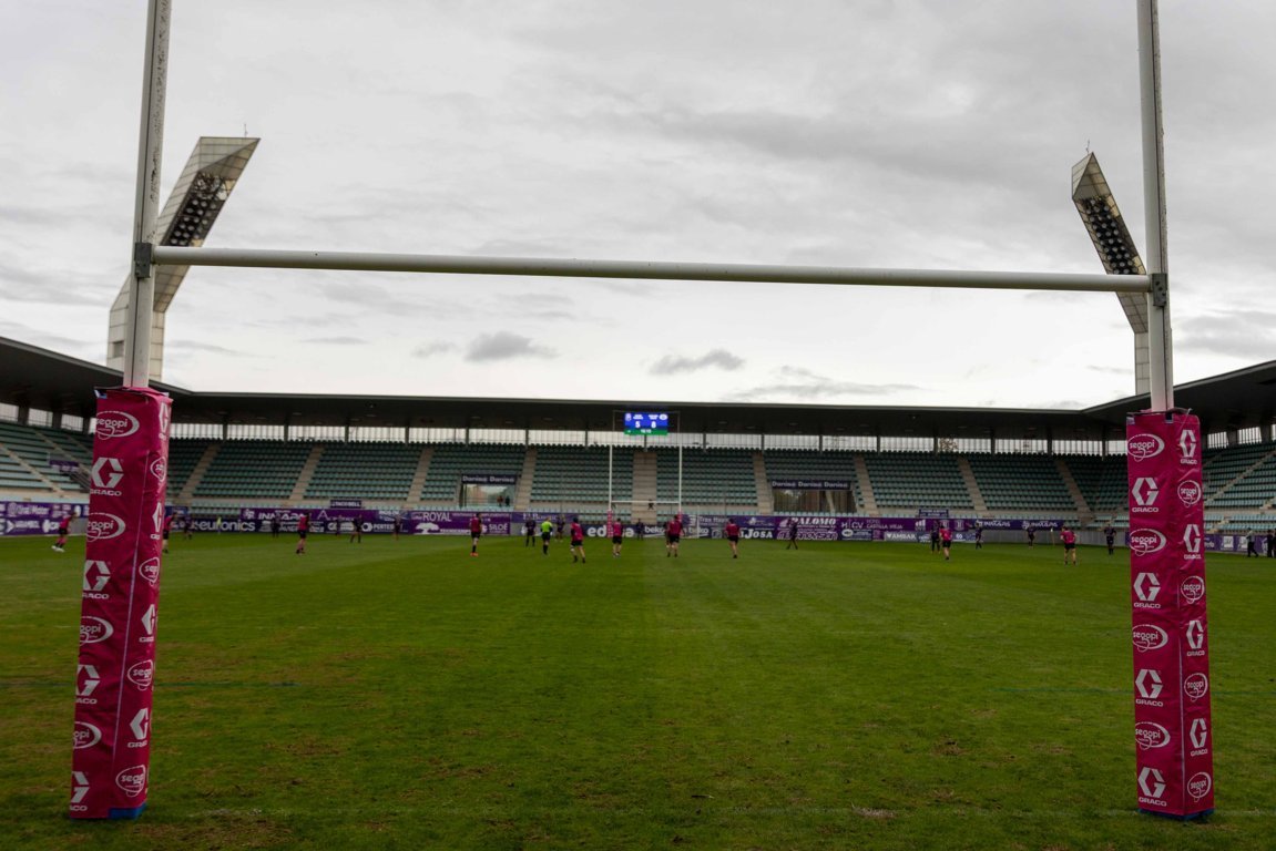 Campo de rugby con jugadores al fondo y postes acolchados en primer plano