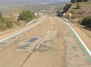 Carretera de dos carriles con asfalto agrietado, parches, señales y guardarraíles.