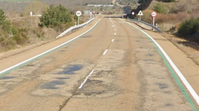 Carretera de dos carriles con asfalto agrietado, parches, señales y guardarraíles.