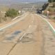 Carretera de dos carriles con asfalto agrietado, parches, señales y guardarraíles.