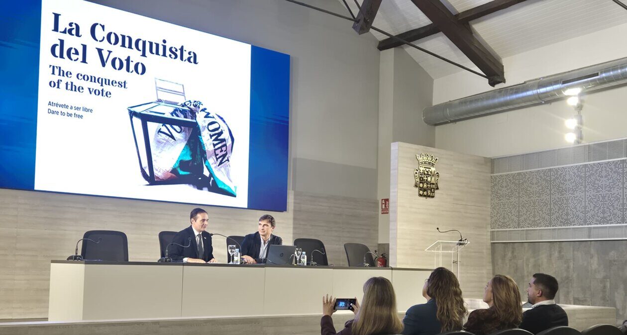 Dos hombres participan en una conferencia ante asistentes y una pantalla sobre el sufragio femenino.