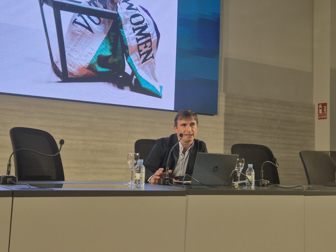 Hombre sentado ante un portátil y un micrófono durante una charla con una proyección detrás.