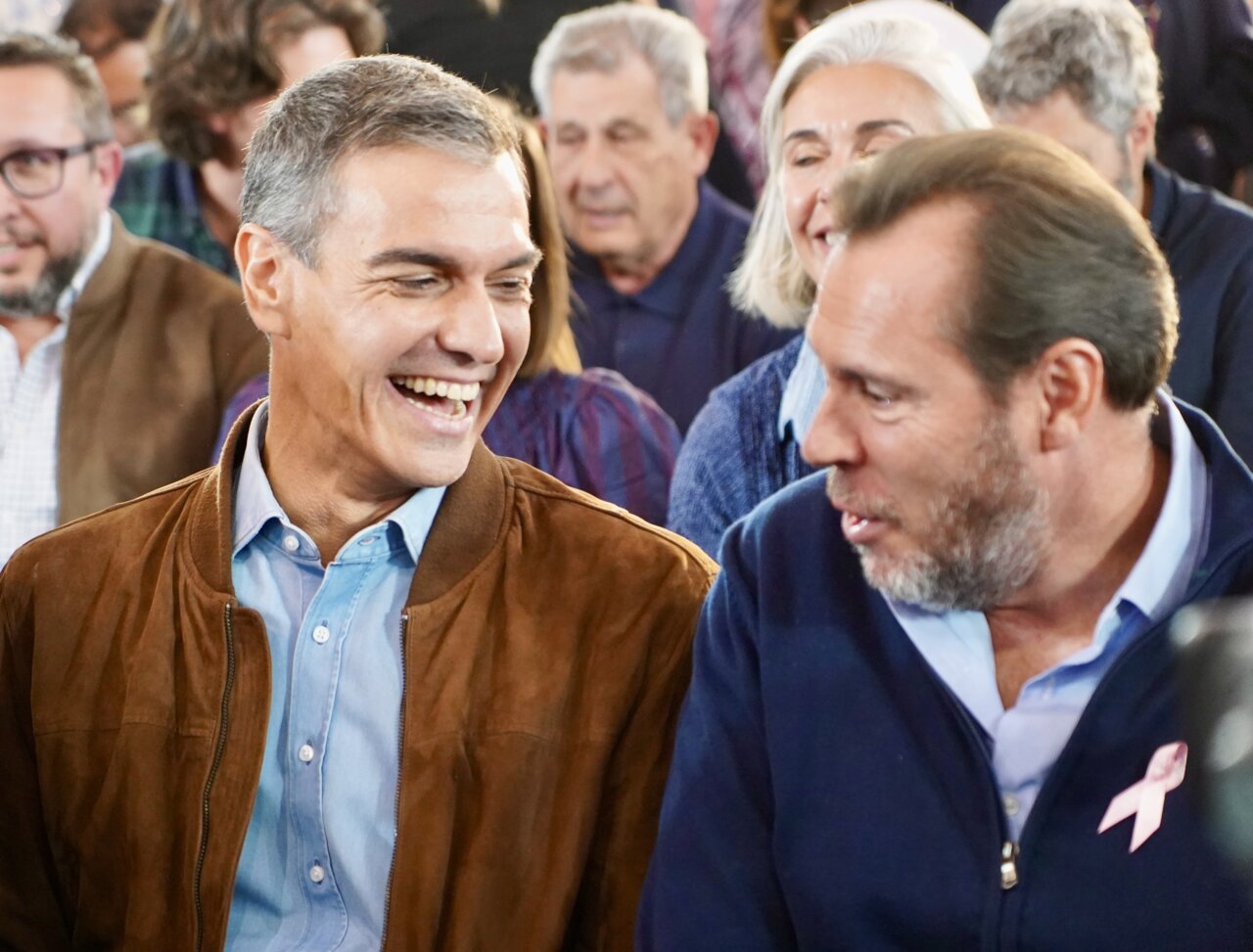 Dos hombres conversan sonrientes entre asistentes reunidos en un espacio interior.