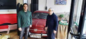 Dos hombres posan junto a un automóvil rojo clásico en un espacio con materiales de tapicería.