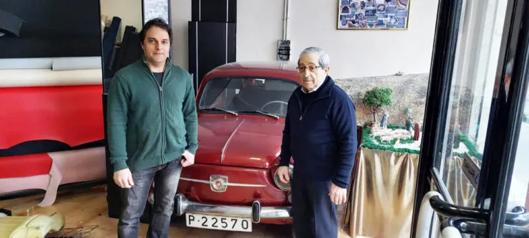 Dos hombres posan junto a un automóvil rojo clásico en un espacio con materiales de tapicería.