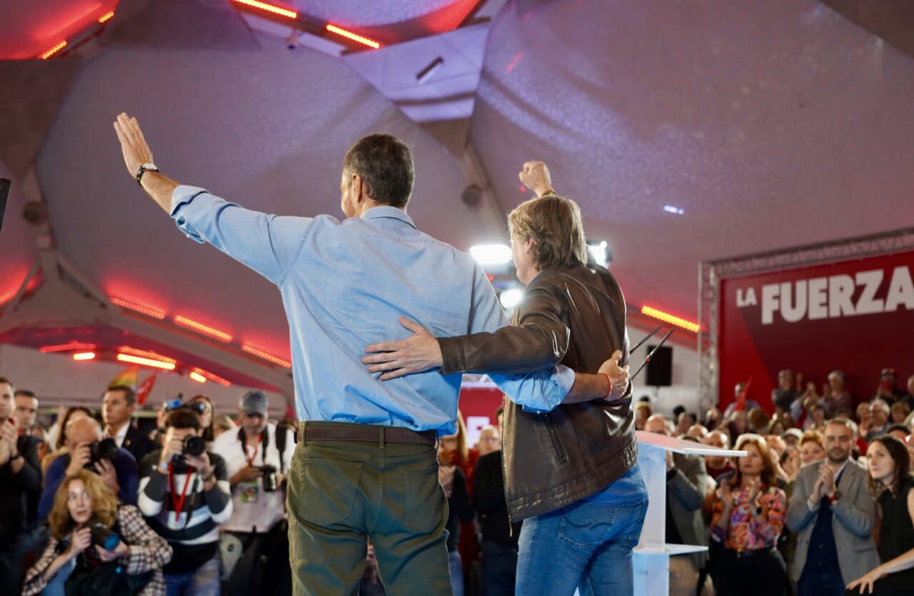 Dos hombres saludan al público desde un escenario mientras los asistentes aplauden.