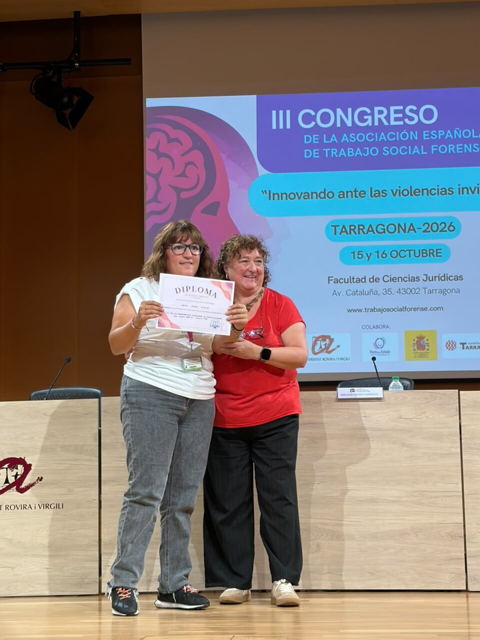 Dos mujeres sostienen un diploma frente a la pantalla del congreso en Tarragona.