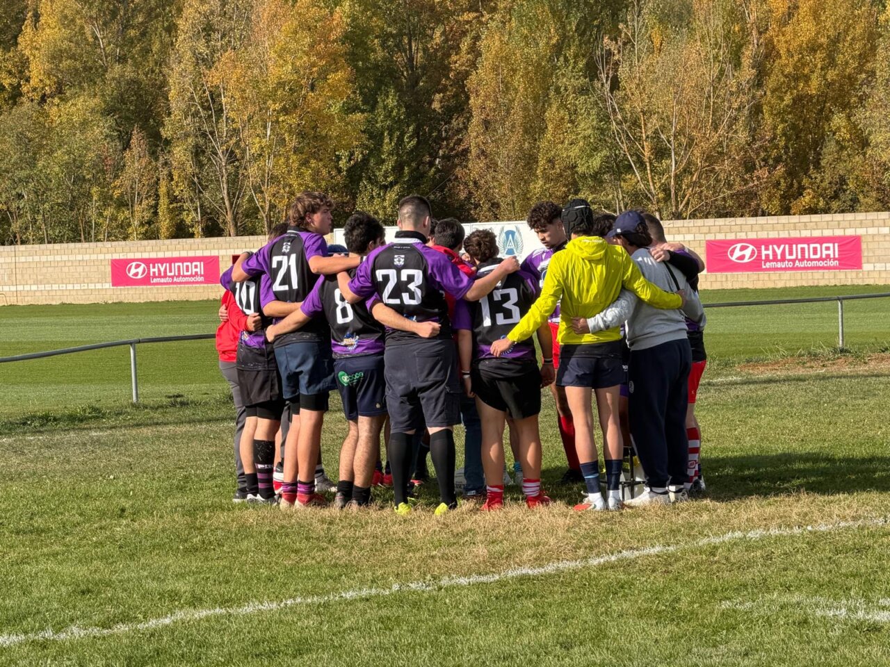Jugadores con camisetas moradas y negras se reúnen en círculo en un campo deportivo