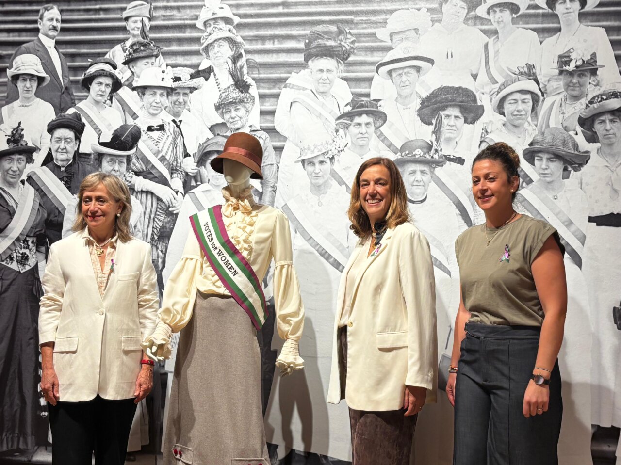 Tres mujeres posando frente a una exposición sobre el sufragio femenino.