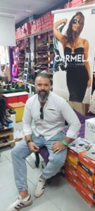 Hombre sentado en una zapatería rodeado de calzado y un cartel de Carmela