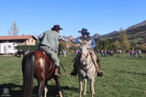Dos jinetes a caballo se saludan en un recinto con más caballos y asistentes.