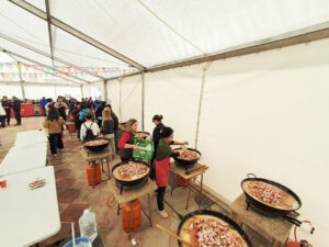 Personas cocinan en grandes sartenes sobre quemadores, bajo una carpa de feria
