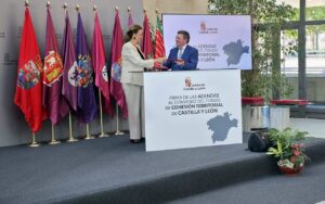 Dos personas intercambian una carpeta ante un atril y varias banderas institucionales.
