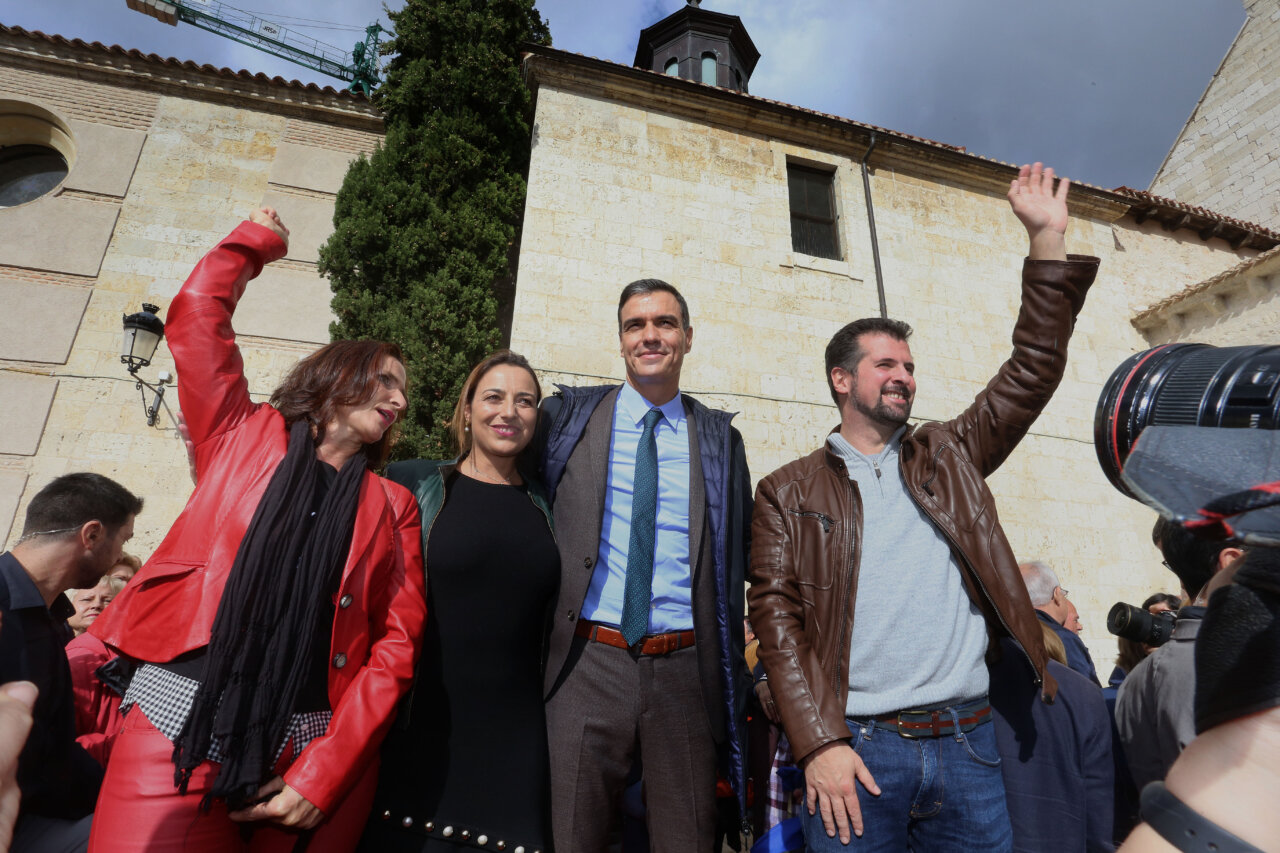 Fotografía de cuatro personas frente a un edificio de piedra; dos levantan la mano.