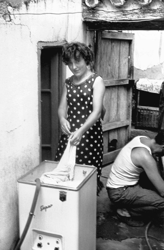 Fotografía en blanco y negro de una mujer junto a una lavadora y un hombre agachado.