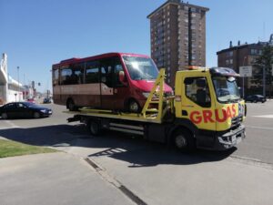 Grúa amarilla de plataforma transporta un autobús rojo por una avenida urbana.