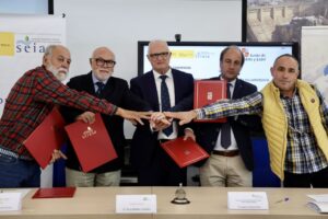 Cinco hombres posan con carpetas rojas y juntan las manos ante carteles institucionales.