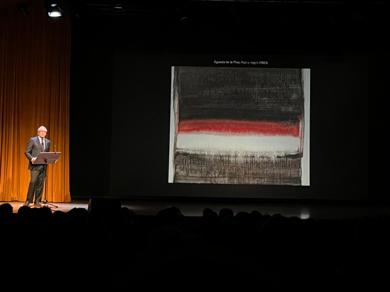 Fotografía de un hombre ante un atril junto a una pintura abstracta proyectada en pantalla.