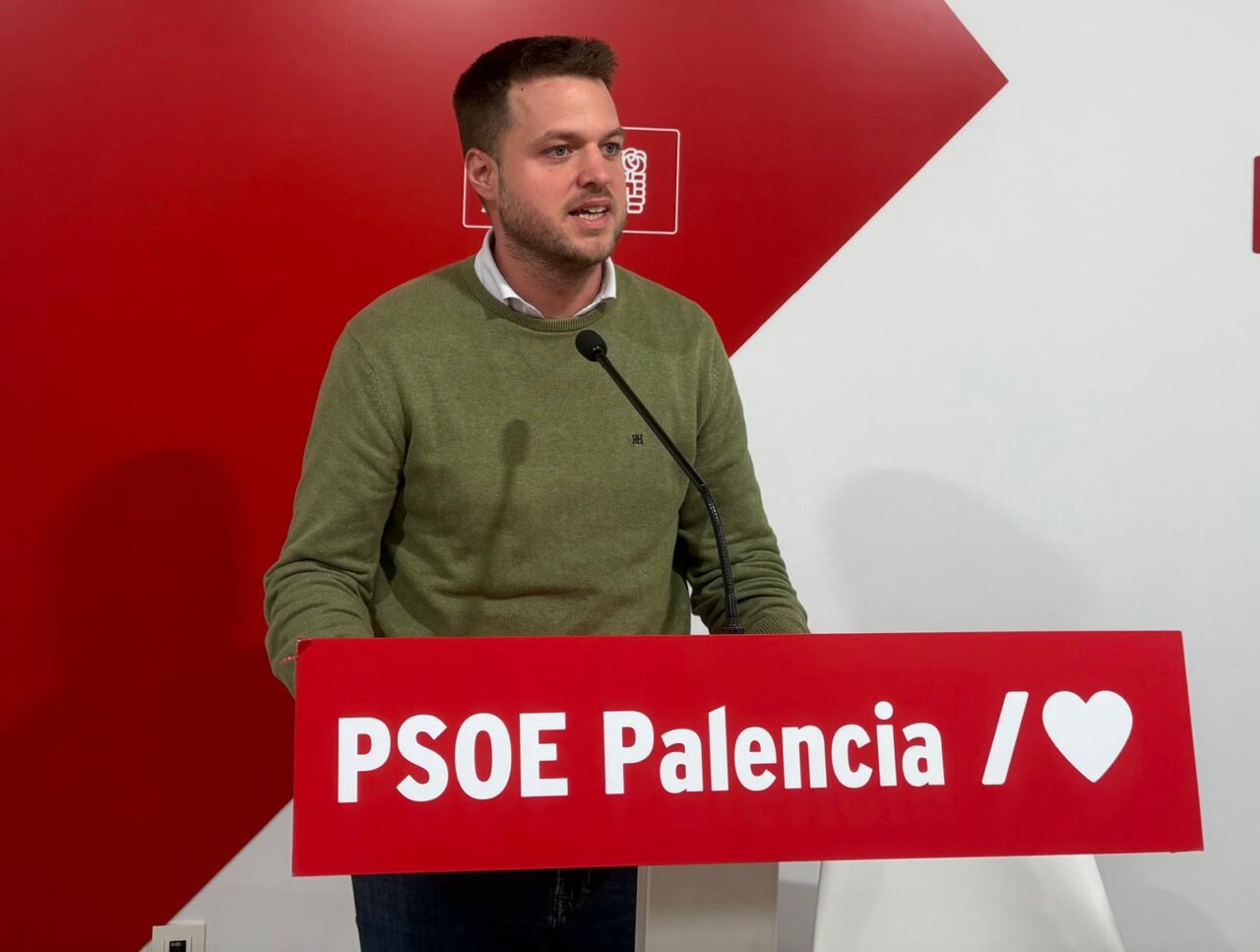 Hombre ante un atril del PSOE Palencia, con micrófono y fondo rojo y blanco