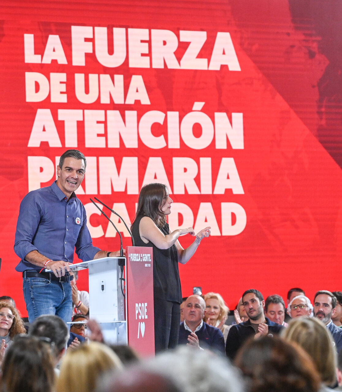 Un hombre habla desde un atril mientras una mujer gesticula ante un público.