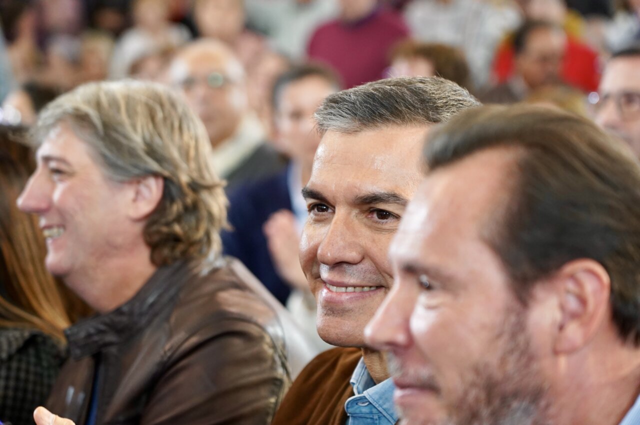 Un hombre sonríe entre varias personas en una reunión multitudinaria bajo techo.