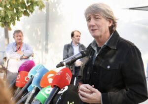 Un hombre habla ante varios micrófonos mientras otras personas observan al fondo.