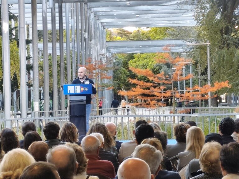 Un hombre habla desde un atril del PP ante un público sentado bajo una estructura metálica.