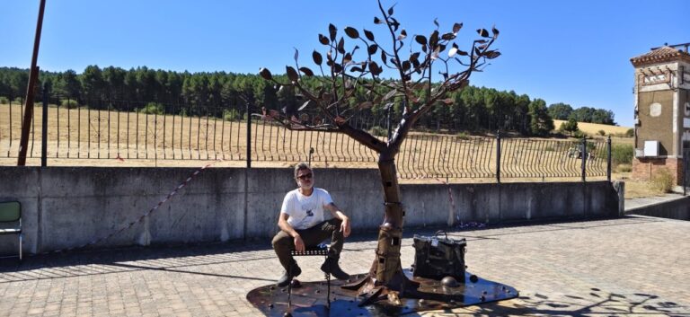 Un hombre sentado junto a una escultura de hierro con forma de árbol.