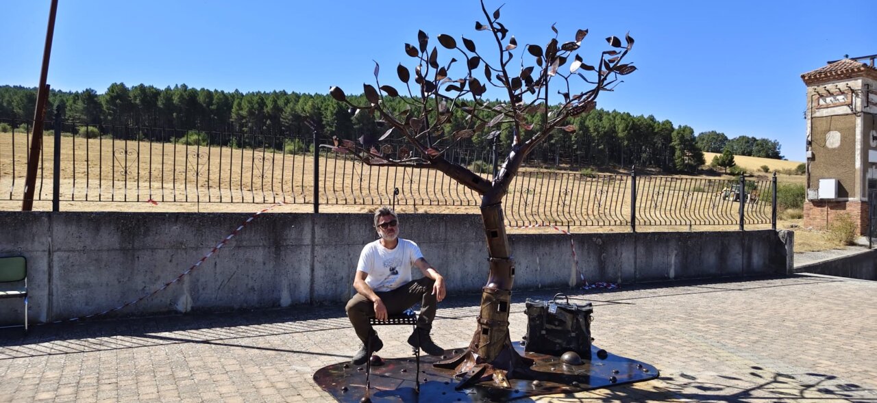 Un hombre sentado junto a una escultura de hierro con forma de árbol.