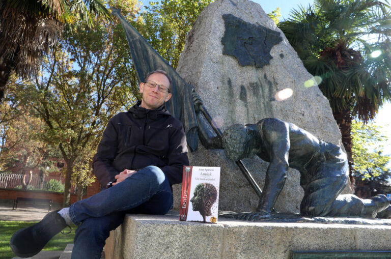 Hombre sentado junto a un libro frente a un monumento escultórico en un parque.