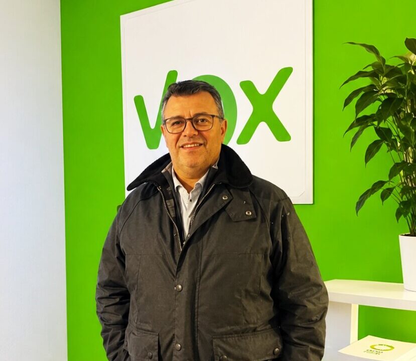 Hombre con gafas y chaqueta oscura posa ante un cartel de VOX.