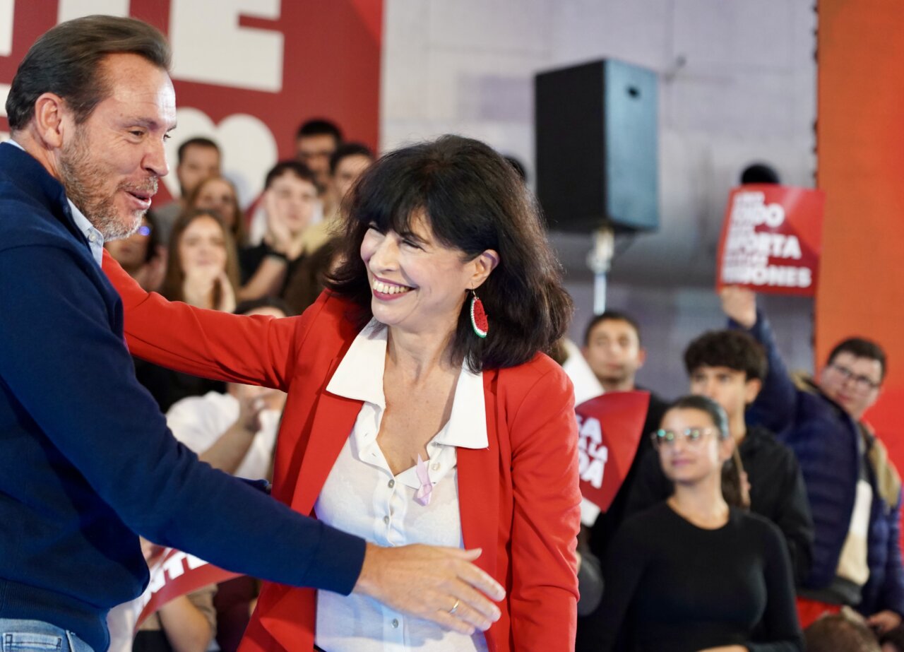 Un hombre y una mujer se saludan frente a asistentes y carteles rojos