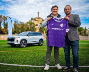 Dos hombres sostienen una camiseta morada JAECOO junto a un SUV en un campo de fútbol