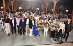 Grupo posa junto a varias mujeres mayores con ramos, ante un público sentado en un salón.